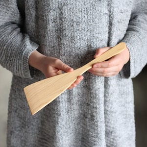 May include: A long, wooden spatula with a wide, flat blade. The spatula is made of light-colored wood and has a smooth, polished finish.