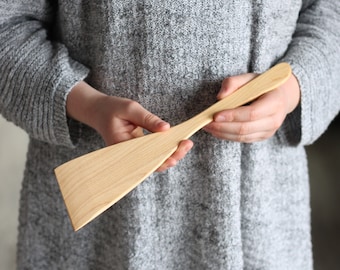 May include: A long, wooden spatula with a wide, flat blade. The spatula is made of light-colored wood and has a smooth, polished finish.