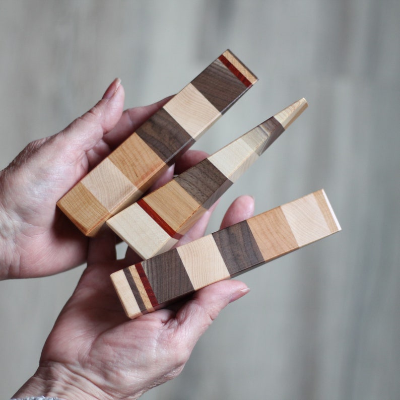 Puede incluir: Tres topes de puerta de madera con un patr&oacute;n de rayas multicolores. Los topes de puerta est&aacute;n hechos de diferentes tipos de madera, incluyendo nogal, arce y cerezo. Los topes de puerta se sostienen en las manos de una persona.