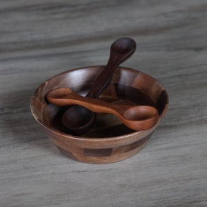 May include: Two wooden spoons in a wooden bowl with a patterned design. The bowl is made of different shades of brown wood.