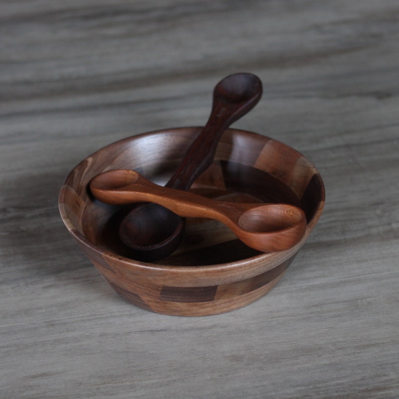 May include: Two wooden spoons in a wooden bowl with a patterned design. The bowl is made of different shades of brown wood.