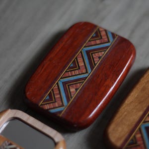 May include: A rectangular wooden box with a geometric inlay design. The box is primarily reddish-brown with a central band of blue, gold, and tan geometric patterns. The box has a glossy finish and appears to be handcrafted.
