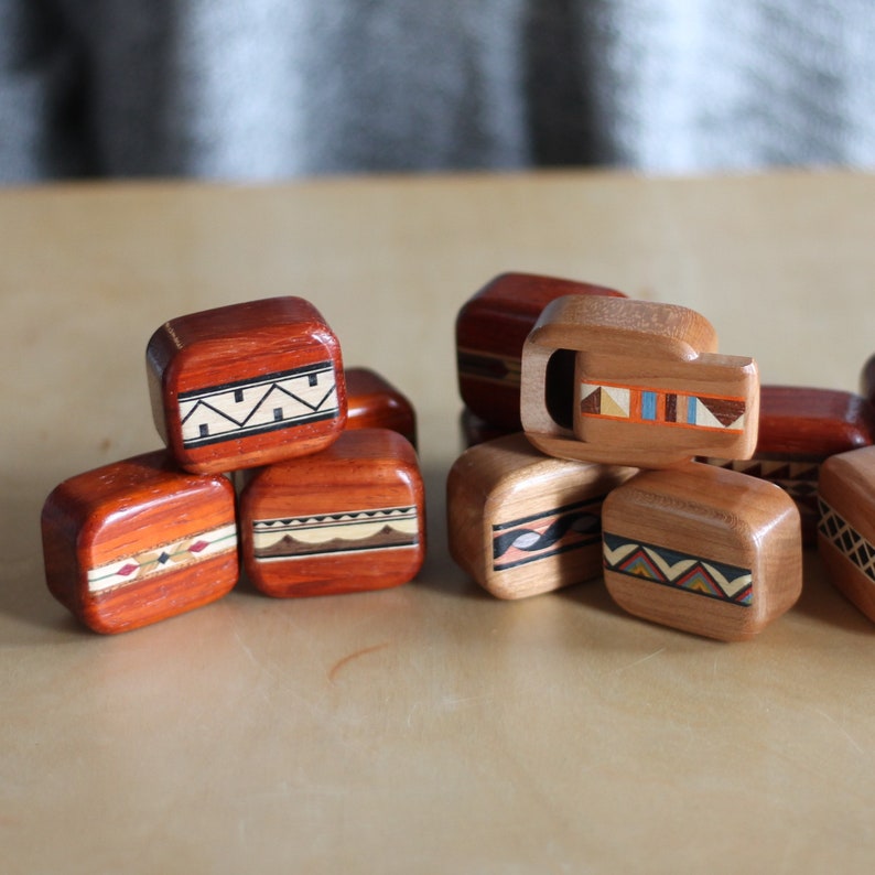 May include: A collection of small, rectangular wooden blocks with rounded edges, featuring intricate inlaid geometric patterns in various colors including black, white, orange, and blue. The blocks are arranged in two clusters, showcasing different wood tones.