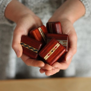 May include: A collection of small, rectangular wooden blocks with a polished reddish-brown finish. Each block features a unique inlaid decorative strip across the center, showcasing various geometric patterns in contrasting light and dark wood tones.