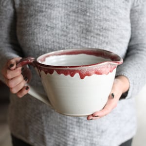 May include: A large white ceramic mixing bowl with a red rim and a handle. The bowl has a speckled glaze and a drip effect around the rim.