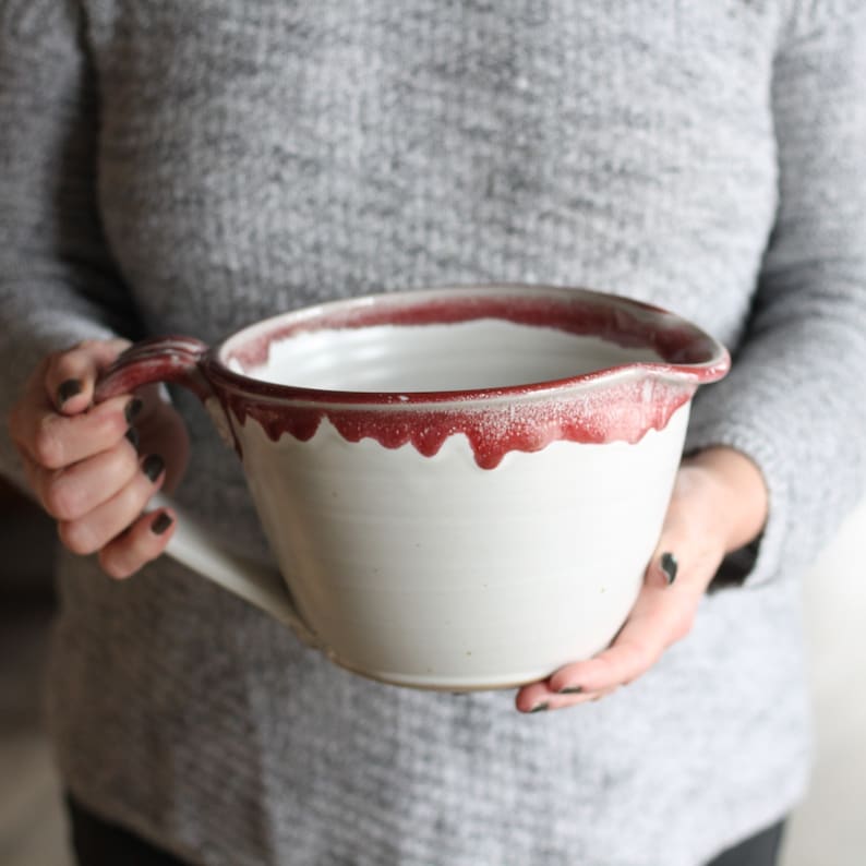 May include: A large white ceramic mixing bowl with a red rim and a handle. The bowl has a speckled glaze and a drip effect around the rim.