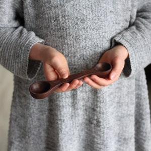 May include: A wooden measuring spoon with two different sized scoops. The spoon is dark brown and has measurements marked on it.
