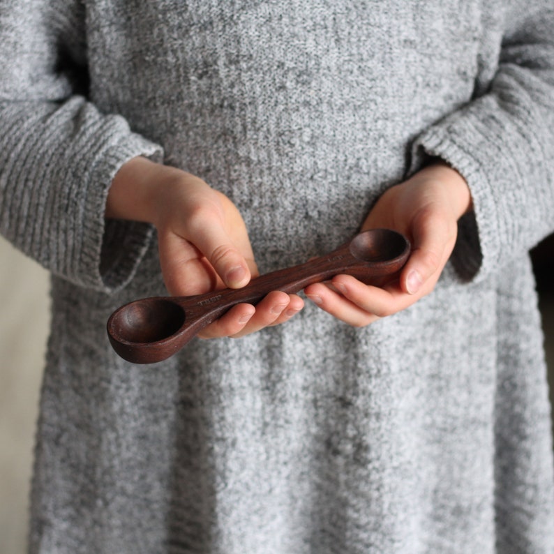 May include: A wooden measuring spoon with two different sized scoops. The spoon is dark brown and has measurements marked on it.