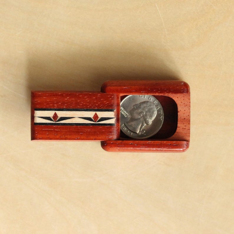 May include: A rectangular reddish-brown wooden sliding box with a decorative inlay strip featuring black, white, and red diamond shapes. The box is partially open, revealing a silver-colored United States quarter coin resting inside the hollow compartment.
