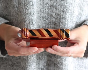 May include: A cylindrical wooden kaleidoscope featuring a diagonal striped pattern in shades of brown, tan, and black, with a clear glass lens at one end. The object is mounted on a rectangular wooden base with a smooth, polished finish.