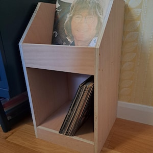 May include: A two-tiered wooden record stand with a light wood finish. The top shelf has a record with a picture of John Lennon on it. The bottom shelf has a stack of records.