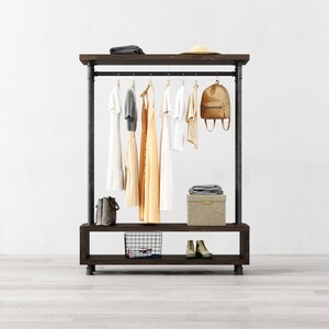 May include: A dark brown wooden clothing rack with a metal pipe rail. The rack has a lower shelf with a wire basket and a top shelf with a straw hat. The rack is holding a variety of clothing items, including dresses, a shirt, and a backpack.