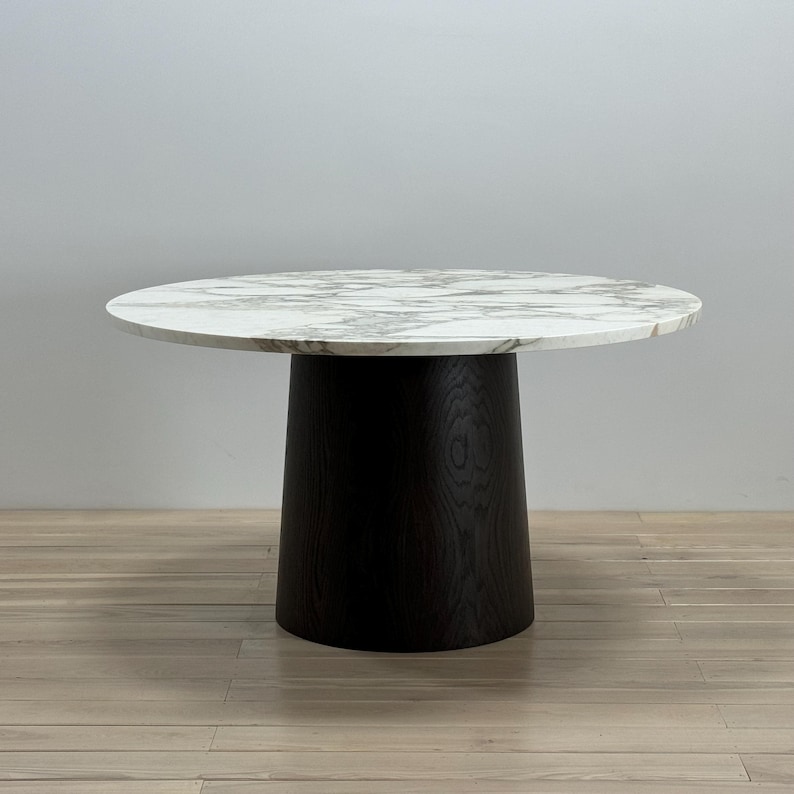 May include: A round marble dining table with a black wood base. The table top has a white marble pattern with grey veining.