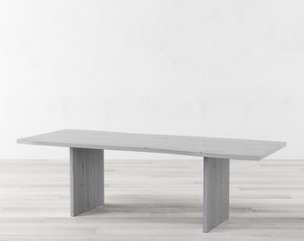 May include: A long, rectangular, whitewashed wood dining table with two rectangular legs on each side. The table has a slightly uneven, organic shape.