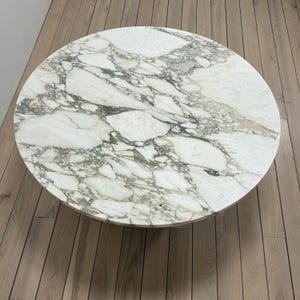 May include: A round marble table top with a white background and grey veining. The table top is sitting on a wooden floor.