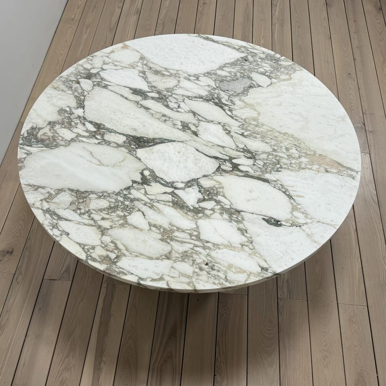 May include: A round marble table top with a white background and grey veining. The table top is sitting on a wooden floor.