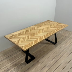 May include: A long, rectangular wooden table with a chevron pattern top and black metal legs. The table is on a wooden floor.