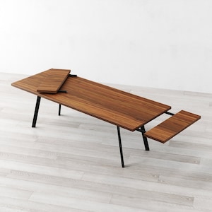 Puede incluir: Una mesa de comedor de madera con patas de metal negro y dos hojas extensibles. La mesa está hecha de madera oscura con un patrón de veta natural. Las hojas están hechas del mismo tipo de madera y están unidas a la mesa con bisagras de metal.