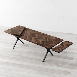 May include: A wooden dining table with a herringbone pattern top and black metal legs. The table has two leaves that can be extended to increase the table's length.