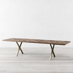 May include: A long, rectangular wooden dining table with a chevron pattern top and gold metal legs. The table is on a light wood floor.