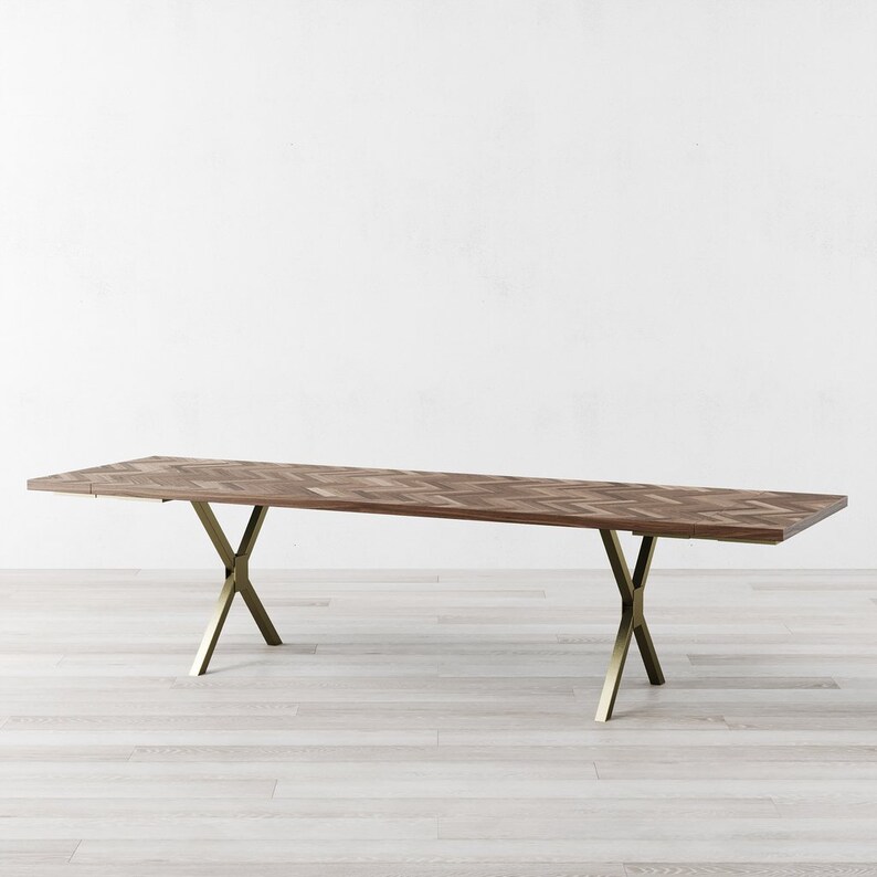 May include: A long, rectangular wooden dining table with a chevron pattern top and gold metal legs. The table is on a light wood floor.