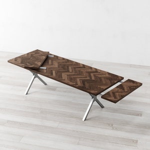 May include: A brown wooden dining table with a herringbone pattern top and silver metal legs. The table has a leaf extension that can be added to the side.