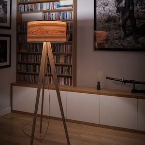 May include: A floor lamp with a cylindrical shade featuring a prominent brown wood grain pattern. The lamp is supported by three slender, light-coloured wooden legs arranged in a tripod configuration, with a thin power cord extending from the base.