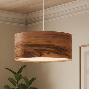 May include: A cylindrical pendant light fitting featuring a natural wood grain veneer shade. The drum-shaped shade is suspended from the ceiling by a thin white cord and a matching white circular canopy, providing downward illumination.