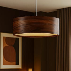 Könnte beinhalten: Eine dunkelbraune, zylindrische Pendelleuchte hängt von der Decke. Das Licht hat eine Holzmaserung und einen warmen, sanften Schein. Die Leuchte hängt über einem Holztisch, auf dem eine Vase und eine Teekanne stehen.