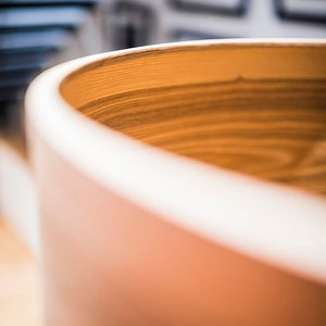 May include: A cylindrical wooden vessel with a smooth, rounded rim and a visible natural grain pattern. The interior of the container displays horizontal wood grain lines in a warm, light brown tone, while the exterior surface is a solid, matte tan.
