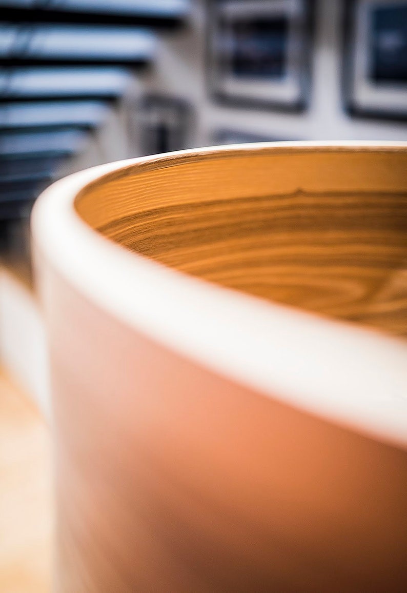 May include: A cylindrical wooden vessel with a smooth, rounded rim and a visible natural grain pattern. The interior of the container displays horizontal wood grain lines in a warm, light brown tone, while the exterior surface is a solid, matte tan.