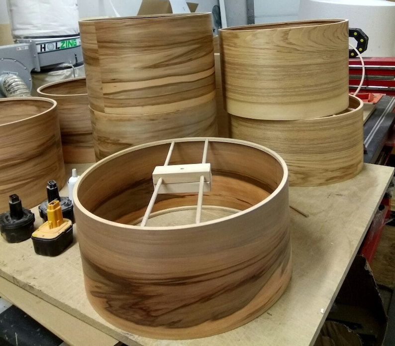 Puede incluir: Primer plano de un casco de tambor de madera en proceso de montaje. El casco est&aacute; hecho de finas tiras de madera que se pegan entre s&iacute; para formar un cilindro. El casco se mantiene unido por un aro de metal y una abrazadera de madera.