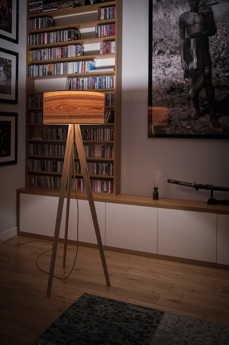 May include: A floor lamp with a cylindrical shade featuring a prominent brown wood grain pattern. The lamp is supported by three slender, light-coloured wooden legs arranged in a tripod configuration, with a thin power cord extending from the base.