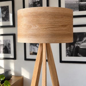 May include: A floor lamp featuring a cylindrical drum lampshade with a light brown wood grain finish, supported by a three-legged tripod base constructed from matching light brown wood with a visible natural grain pattern throughout the structure.