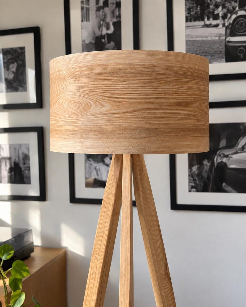 May include: A floor lamp featuring a cylindrical drum lampshade with a light brown wood grain finish, supported by a three-legged tripod base constructed from matching light brown wood with a visible natural grain pattern throughout the structure.
