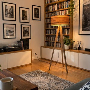 May include: A tripod floor lamp with a cylindrical wooden shade featuring a prominent grain pattern. The lamp stands on three slender wooden legs and emits a warm, soft glow, illuminating the surrounding space in a living room setting.