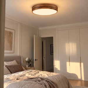 May include: A round flush mount ceiling light fixture featuring a circular wooden frame with a visible grain pattern and a flat, white, translucent diffuser that emits a warm, soft glow, illuminating the room from the center of the ceiling.