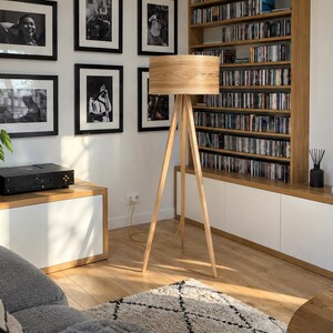 May include: A floor lamp with a cylindrical light-coloured wood veneer shade and a three-legged tripod base made of matching light wood. The lamp stands upright on a wooden floor, featuring a simple design with a visible power cord extending downwards.