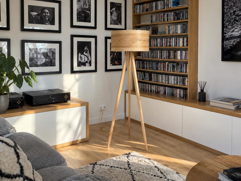 May include: A floor lamp with a cylindrical light-coloured wood veneer shade and a three-legged tripod base made of matching light wood. The lamp stands upright on a wooden floor, featuring a simple design with a visible power cord extending downwards.