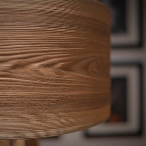 May include: A cylindrical lamp shade featuring a textured light brown surface with a detailed wood grain pattern. The material displays horizontal ridges and grooves that mimic the natural appearance of wood, creating a tactile finish.