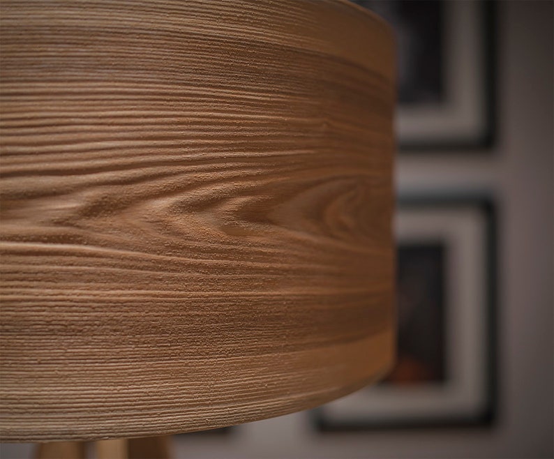 May include: A cylindrical lamp shade featuring a textured light brown surface with a detailed wood grain pattern. The material displays horizontal ridges and grooves that mimic the natural appearance of wood, creating a tactile finish.