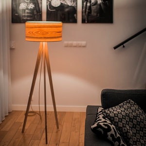May include: A floor lamp with a tripod base made of light-coloured wood and a cylindrical drum shade featuring a prominent orange and brown wood grain pattern. The lamp emits a warm, soft glow, illuminating the surrounding area from within.