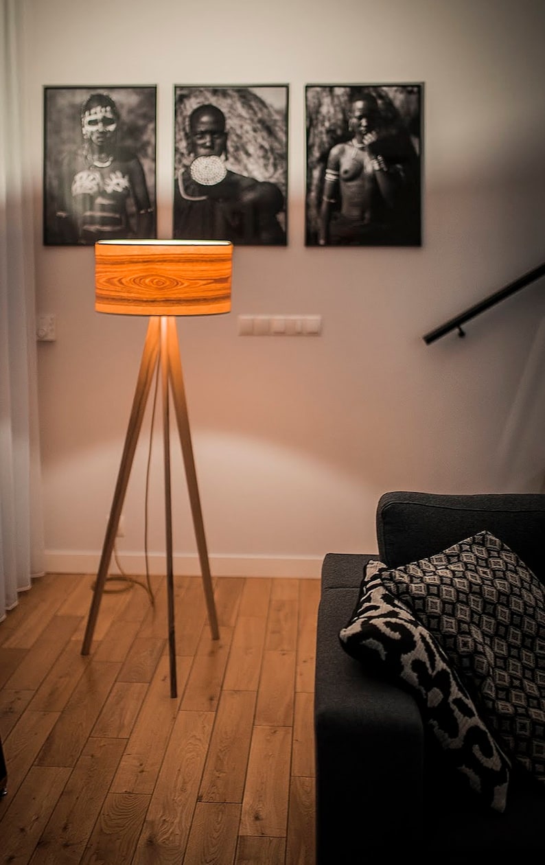 May include: A floor lamp with a tripod base made of light-coloured wood and a cylindrical drum shade featuring a prominent orange and brown wood grain pattern. The lamp emits a warm, soft glow, illuminating the surrounding area from within.