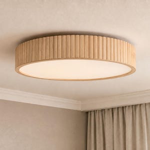 May include: A round flush mount ceiling light fixture featuring a light-toned wood frame with a vertical fluted or ribbed texture around the exterior edge and a flat, circular white diffuser panel centered on the underside of the fixture.