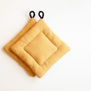 Può includere: Due presine trapuntate giallo senape con anelli neri per appenderle. Le presine sono quadrate con un quadrato più piccolo al centro. Il tessuto ha un aspetto strutturato. Le presine sono su sfondo bianco.