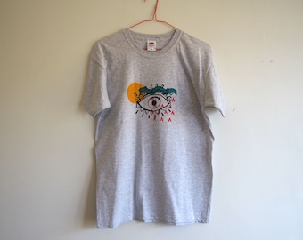 May include: Gray t-shirt with a colorful eye graphic. The eye is outlined in black and has a green eyelid, a yellow sun behind it, and red teardrop shapes falling from it.