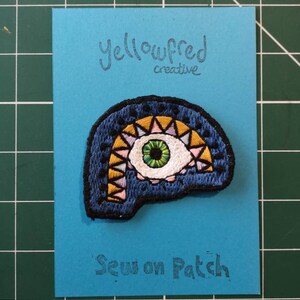 May include: A blue, yellow, pink, and green embroidered eye patch with the text "Sew on Patch" and "Yellowfred creative" on a blue background.