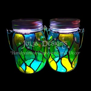 May include: Two glass jars with solar-powered lights inside. The jars are hand-painted with a stained glass design in shades of green, blue, and yellow. The text "Julia Designs" and "Handpainted Glassware and Decor" is visible on the jars.