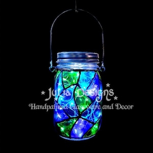 以下が含まれることがあります： 金属製のハンドルと蓋が付いた、手描きのガラス瓶。 青と緑の色合いのステンドグラスのデザインで、小さなライトで照らされています。 「Julia Designs Handpainted Glassware and Decor」の文字が見えます。