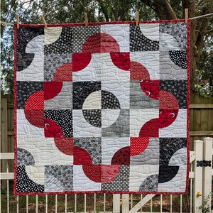 May include: A handmade quilt with a geometric design, featuring red, black, white, and gray fabrics. The quilt is square, with a red border and a pattern of curved shapes and squares. It is hanging outside on a clothesline.